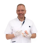 Zubní hygienista Simon Kvapil v bílém plášti se usmívá a drží dentální nástroje s modelem čelisti.